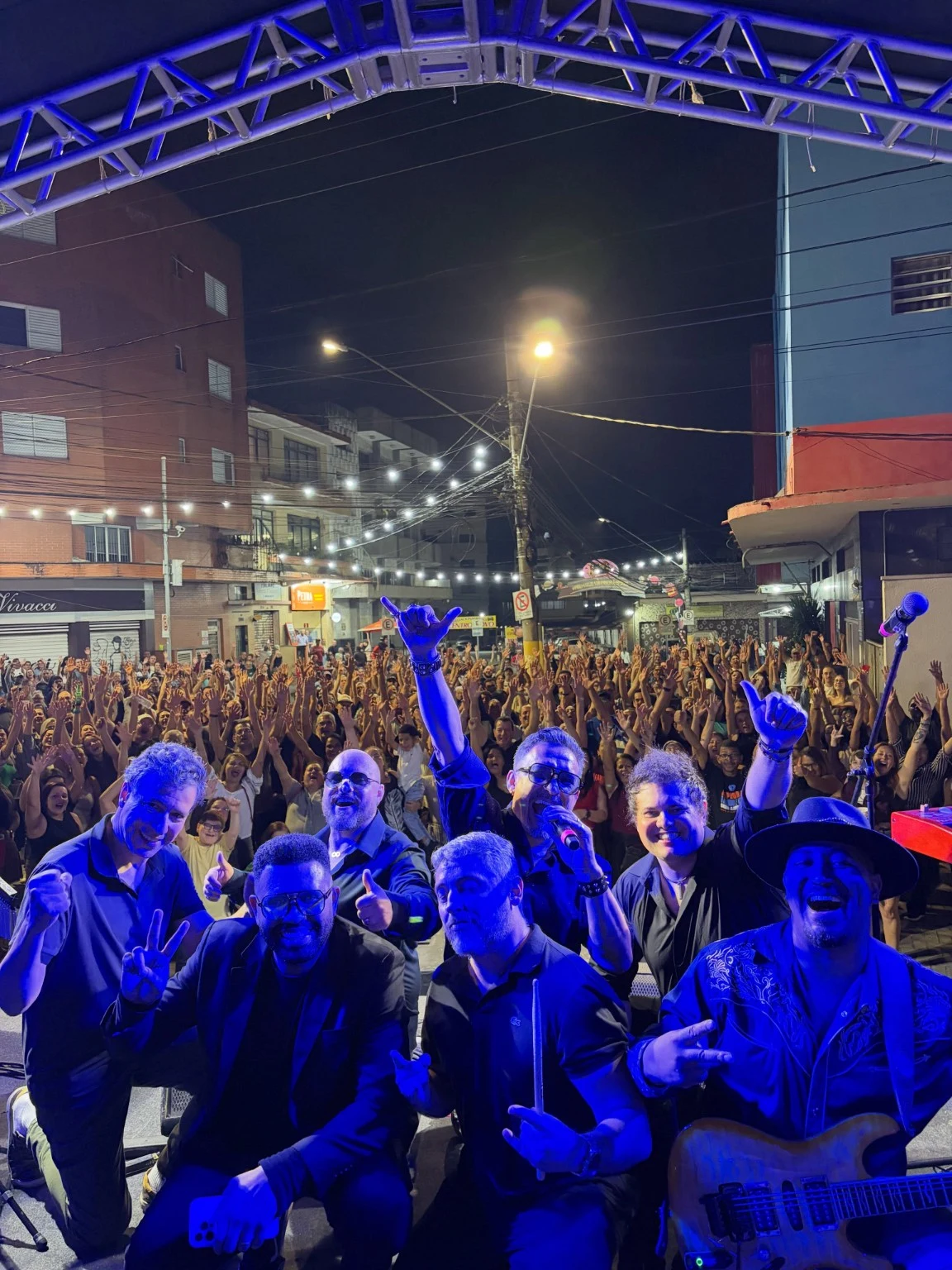 Integrantes da banda diante do público em um show ao ar livre, sob iluminação azul
